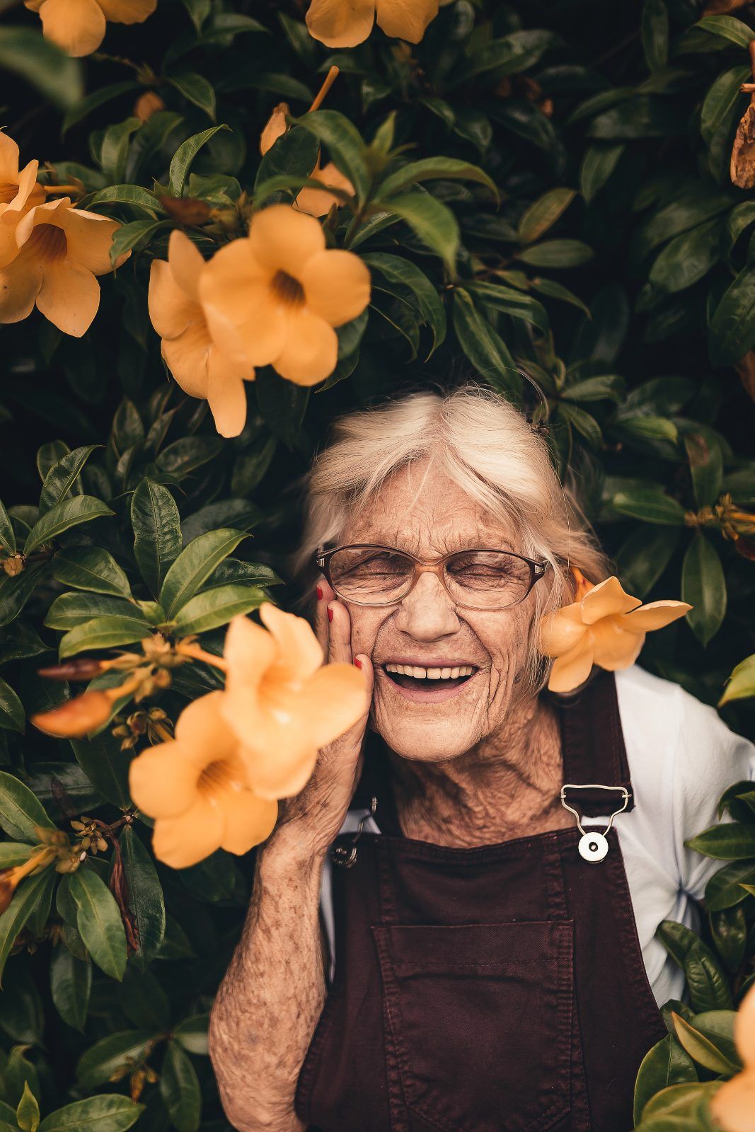 Older woman in glasses laughing among yellow flowers in a garden
